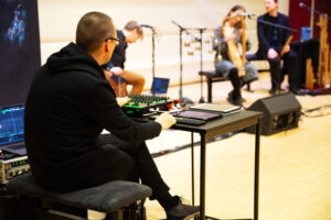 Kuvassa mies, jonka selkä kameraan päin, joka istuu äänitekniikkapöydän ääressä. Taustalla on yhtye lavalla.